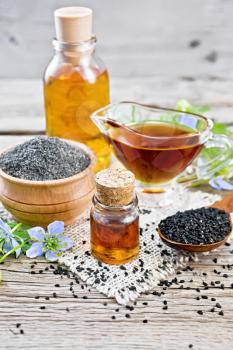 Nigella sativa oil in vial, gravy boat and bottle, seeds in a spoon and black cumin flour in a bowl on burlap, kalingi twigs with blue flowers and green leaves on background of an old wooden board