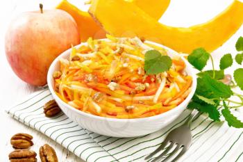 Pumpkin, carrot and apple salad with pecans seasoned with vegetable oil in a bowl on a towel, mint on white wooden board background