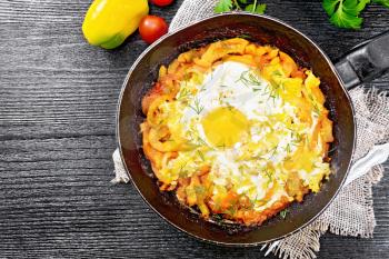 Fried eggs with tomatoes, sweet pepper, onions and herbs in a pan on burlap, parsley, fork on wooden board background from above