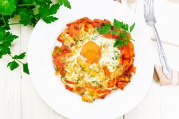 Fried eggs with tomatoes, sweet pepper, onions and herbs in a plate, napkin, parsley and fork on wooden board background from above