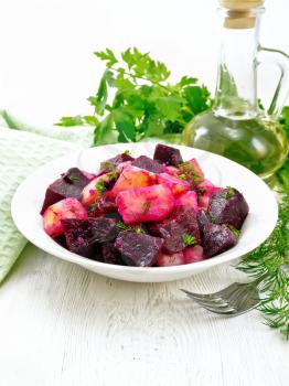 Beetroot and potato salad, seasoned with vegetable oil and vinegar in a plate, napkin, parsley and dill on a white wooden board background