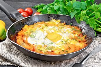 Fried eggs with tomatoes, sweet pepper, onions and herbs in a pan on burlap, parsley, fork on wooden board background