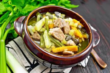 Eintopf soup of pork, celery, beans, carrots and potatoes with leek in a clay bowl on a towel on wooden board background