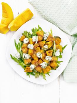Salad of baked pumpkin, arugula with balls of salt cheese, seasoned with honey, grainy mustard, garlic and vegetable oil in a plate, napkin and fork on background of light wooden board from above
