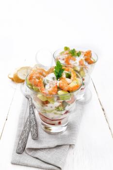 Puff salad with shrimp, avocado, fresh cucumber, sweet pepper and tomato, seasoned with yogurt sauce in two glass glasses on a towel, bread and forks on a wooden board background

