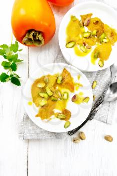 Dessert of yogurt, persimmon and honey with vanilla, cardamom and pistachios in two bowls on a napkin, mint and spoon on the background of light wooden board on top