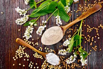 Buckwheat flour from brown and green cereals in two spoons, flowers and leaves on the background of wooden board from above
