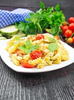 Fusilli with chicken, zucchini and tomatoes in a plate, napkin, fork, basil and parsley on dark wooden board background