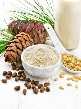 Cedar flour in a bowl, nuts and cones, spoon with peeled nuts, green pine branch and cedar milk in bottle on white wooden board background