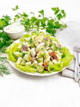 Salad from salmon, cucumber, eggs and avocado with mayonnaise on lettuce leaves in a plate, napkin, dill, parsley and fork on a wooden board background