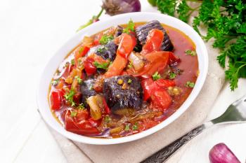 Vegetable ragout with eggplant, tomatoes, bell peppers, onions and spices in a plate on kitchen towel, garlic, parsley, hot peppers and a fork on wooden board background