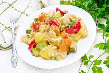 Vegetable ragout with zucchini, cabbage, potatoes, tomatoes and bell peppers in creamy sauce in plate, a towel, parsley and a fork on wooden board background