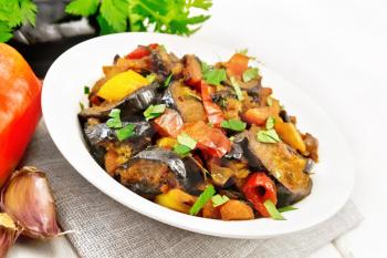 Vegetable ragout with eggplant, tomatoes, sweet and hot peppers, onions, carrots, fried with herbs and spices in plate on a napkin, garlic, parsley on white wooden board background