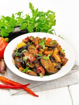 Vegetable ragout with eggplant, tomatoes, sweet and hot peppers, onions, carrots, fried with herbs and spices in plate on a towel, garlic, parsley on white wooden board background