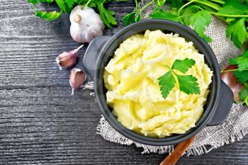 Mashed potatoes in a black saucepan and a spoon on burlap, garlic, parsley on wooden board background from above