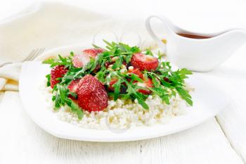 Strawberry, couscous, cedar nuts and arugula salad dressed with balsamic vinegar and olive oil in plate, a napkin and a fork on light wooden board background