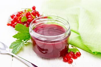 Red currant jam in glass jar, bunches of berries with leaves, a napkin and a spoon on light background of a wooden board