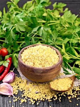Bulgur groats - steamed wheat grains - in a clay bowl and a spoon on burlap napkin, tomatoes, hot peppers, garlic and parsley on wooden board background