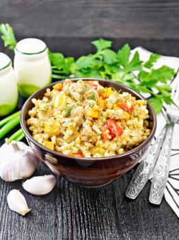 Barley porridge with minced meat, yellow and red bell peppers, garlic and onions in a clay bowl, napkin and parsley on dark wooden board background