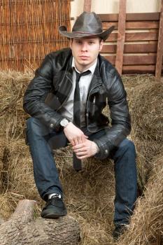 A young cowboy in leather jacket in the rural interior is sitting on a haystack.