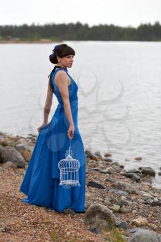 Girl in a blue dress with an empty bird cage stands in cloudy, rainy weather on the lake.
