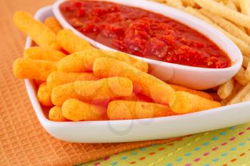 Potato, corn chips in bowl and red sauce on colorful tablecloth.