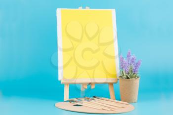 Wooden easel with blank canvas, brushes and lavender flowers on blue background.
