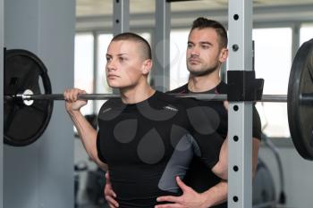 Personal Trainer Showing Young Man How To Train Barbell Squats Exercise In A Health And Fitness Concept