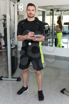 Personal Trainer With Clipboard Monitors People While They Exercise In A Gym Or Fitness Club