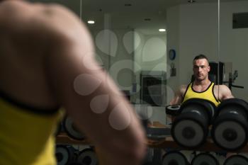 Young Strong Man In The Gym And Exercising Shoulders With Dumbbells At Mirror - Muscular Athletic Bodybuilder Fitness Model Exercise Shoulder