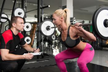Young Woman Exercise Legs Squat  In The Gym While Personal Trainer Helps Out