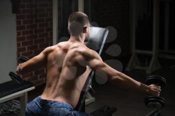 Handsome Man Working Out Back And Shoulders With Dumbbells In A Dark Gym