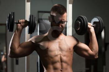 Handsome Man Wearing Eyeglasses Working Out Shoulders With Dumbbells In A Dark Gym