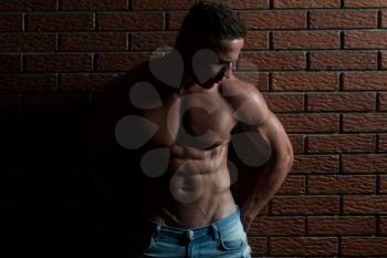 Healthy Young Man Standing Strong In The Gym And Flexing Muscles - Muscular Athletic Bodybuilder Fitness Model Posing After Exercises On Wall of Bricks
