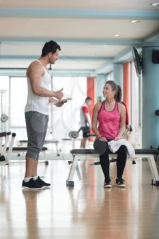 Personal Trainer Takes Notes While Young Woman Resting On Bench In Gym