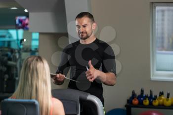 Personal Trainer Showing Ok Sign To Client - Young Woman Exercise Legs On Machine In The Gym Or Fitness Club