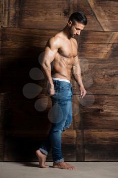 Healthy Young Man Standing Strong Against a Wooden Wall and Flexing Muscles While Wearing Blue Jeans - Muscular Athletic Bodybuilder Fitness Model Posing After Exercises - a Place for Your Text
