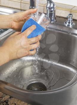 Vertical photo of female hands cleaning a drinking glass with water running from kitchen faucet sink