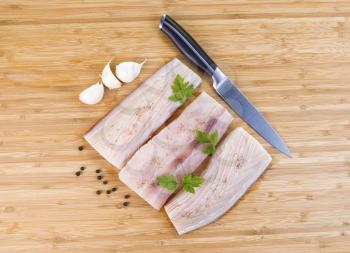 Horizontal photo of white fish fillets, knife, parsley, garlic and pepper salts on Natural Bamboo Wood background