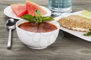 Fresh tomato and basil soup with melon, tuna fish sandwich, cucumber and glass of water on aging wood as background
