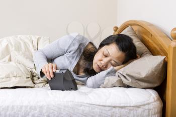 Horizontal photo of mature woman, lying head down in pillow, while holding alarm clock in other hand