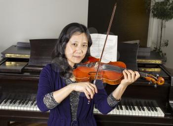 Mature Asian woman playing violin with old dark wooden piano in background