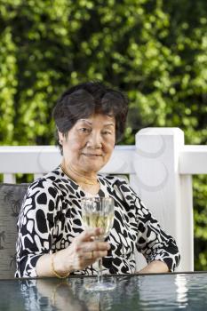Vertical photo of senior Asian woman drinking white wine on a nice day with white patio railing and plush green trees in the background