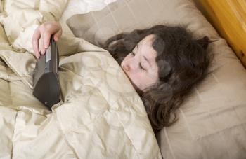 Horizontal photo of young girl looking at alarm clock in surprise, while in bed
