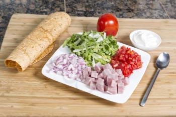 Horizontal photo of sliced tomato, lettuce, onion and ham in white plate with sour cream in small bowl, spoon, and soft taco shell on natural bamboo cutting board