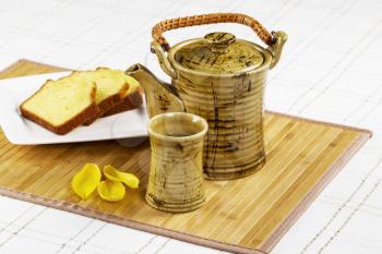 Horizontal photo of ceramic tea pot with cup, Golden Lemon Pound Cake, yellow leafs on bamboo place mat with White Striped Table Cloth in background