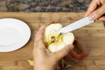 Horizontal photo of female hands removing pit of freshly peeled apple using a paring knife with bamboo cutting board, white dish and stone counter top in background