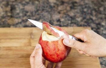 Horizontal photo of female hands peeling skin off of apple using a paring knife with bamboo cutting board and stone counter top in background