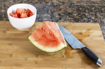 Horizontal photo of freshly cut watermelon with large kitchen knife, white ceramic bowl and bamboo cutting board on granite stone counter top 