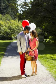 Vertical photo of young adult couple dressed in formal attire looking at each other while holding several balloons, picnic basket with walking path, green grass and trees behind them  